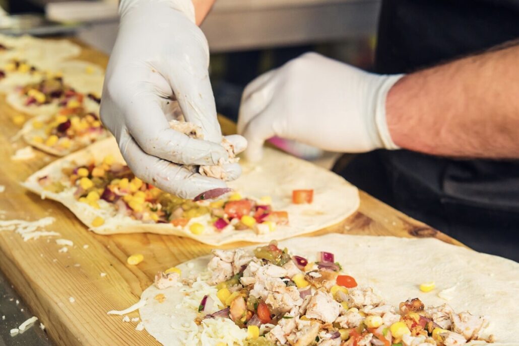 Hände in Handschuhen belegen Tortillas mit Hühnerfleisch, Mais und Tomaten am Street Food Festival