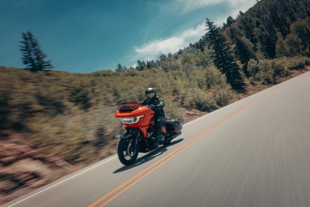 Zu sehen ist ein orangefarbenes Touring-Motorrad in Dreiviertelansicht vorne links während der Fahrt auf einer asphaltierten Bergstraße, gesäumt von bewaldeten Hängen.