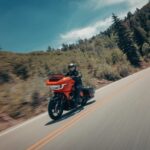 Zu sehen ist ein orangefarbenes Touring-Motorrad in Dreiviertelansicht vorne links während der Fahrt auf einer asphaltierten Bergstraße, gesäumt von bewaldeten Hängen.