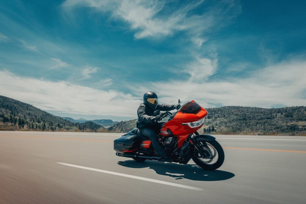 Ein rot-schwarzes Harley-Davidson-Touring-Motorrad fährt in Dreiviertelansicht vorne rechts auf einer asphaltierten Straße vor einer hügeligen Landschaft unter blauem Himmel.