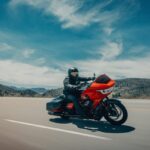Ein rot-schwarzes Harley-Davidson-Touring-Motorrad fährt in Dreiviertelansicht vorne rechts auf einer asphaltierten Straße vor einer hügeligen Landschaft unter blauem Himmel.