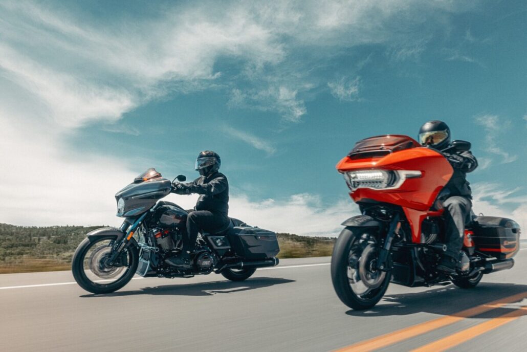 Zwei schwere Touring-Motorräder, links ein dunkles und rechts ein orangenfarbenes Modell, in Seitenansicht und Dreiviertelansicht während der Fahrt auf einer asphaltierten Landstraße unter blauem Himmel.