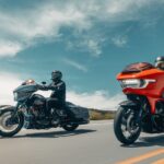 Zwei schwere Touring-Motorräder, links ein dunkles und rechts ein orangenfarbenes Modell, in Seitenansicht und Dreiviertelansicht während der Fahrt auf einer asphaltierten Landstraße unter blauem Himmel.